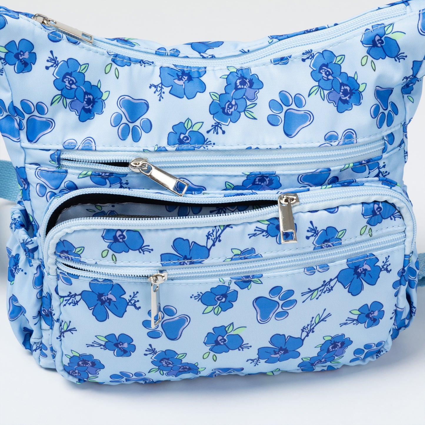 A light blue crossbody bag with a medium blue floral pattern, featuring multiple zippered pockets and an adjustable shoulder strap, shown against a plain white background.