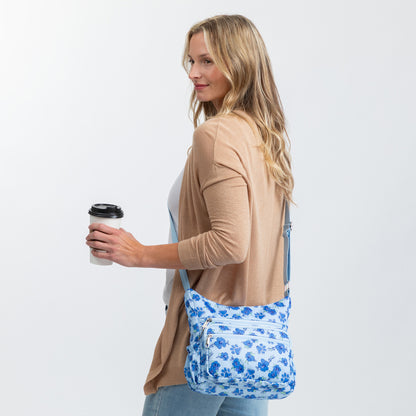 A woman in a tan cardigan and jeans carries a blue and white floral and paw print patterned crossbody bag while holding a coffee cup against a plain light gray background.