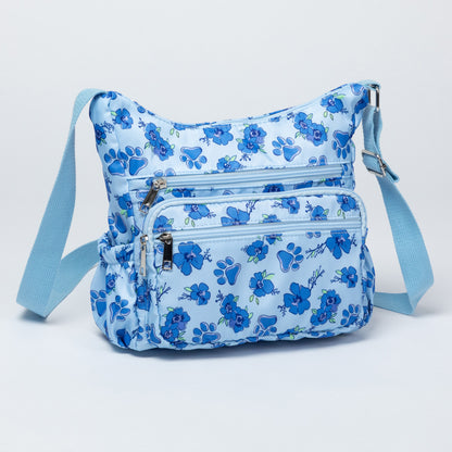 A light blue crossbody bag with a medium blue floral pattern, featuring multiple zippered pockets and an adjustable shoulder strap, shown against a plain white background.