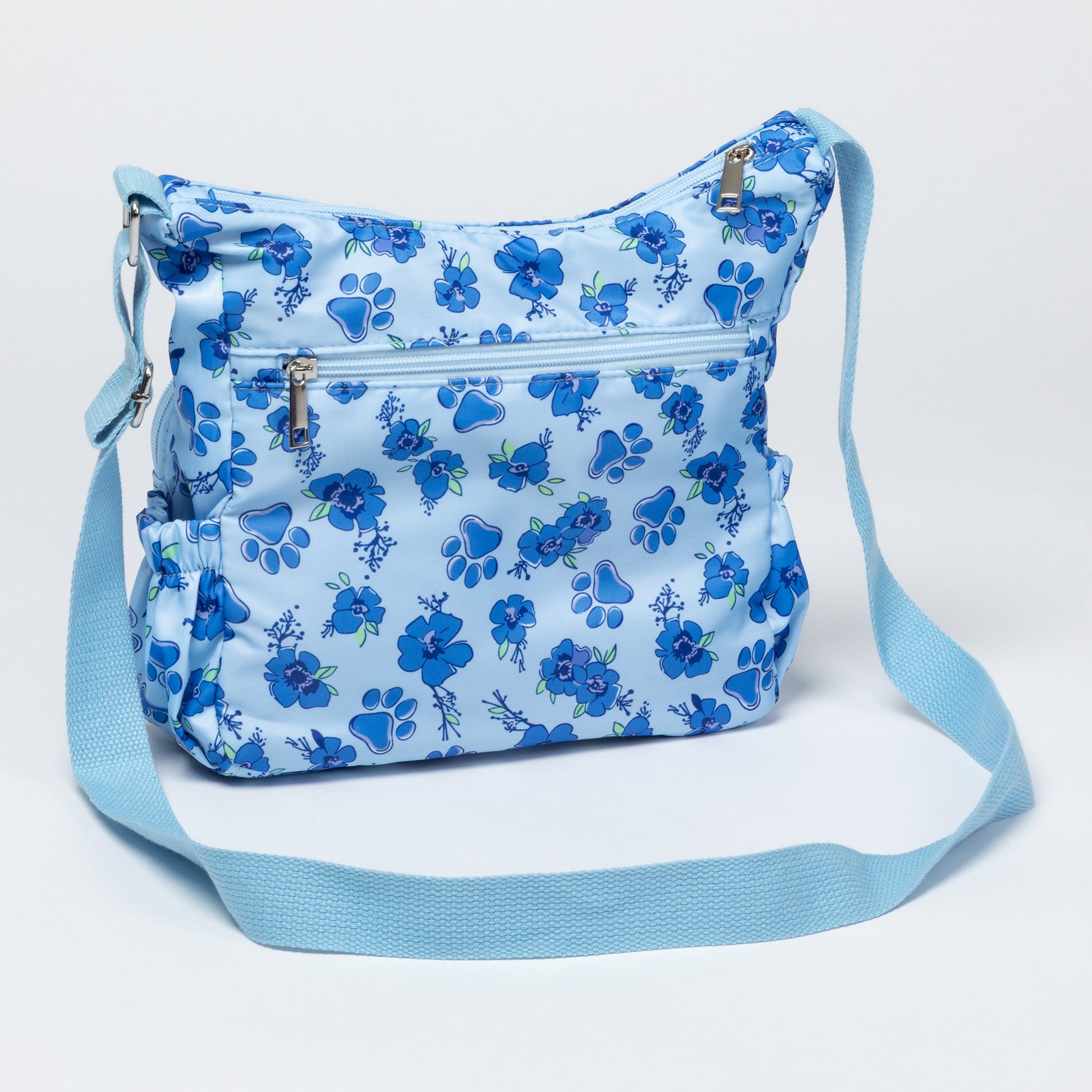 A light blue crossbody bag with a medium blue floral pattern, featuring multiple zippered pockets and an adjustable shoulder strap, shown against a plain white background.