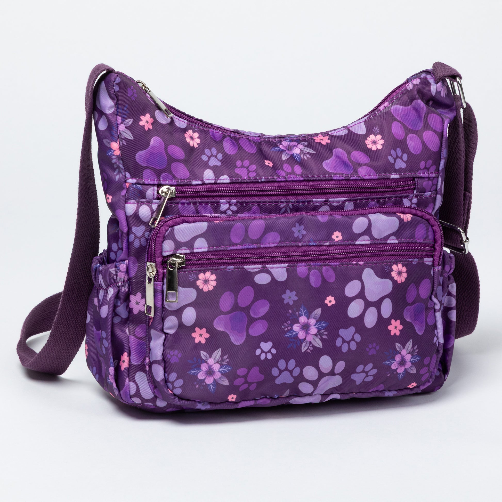 A purple crossbody bag with a floral and paw-print pattern, featuring multiple zippered pockets and a matching adjustable shoulder strap, shown on a light gray background.