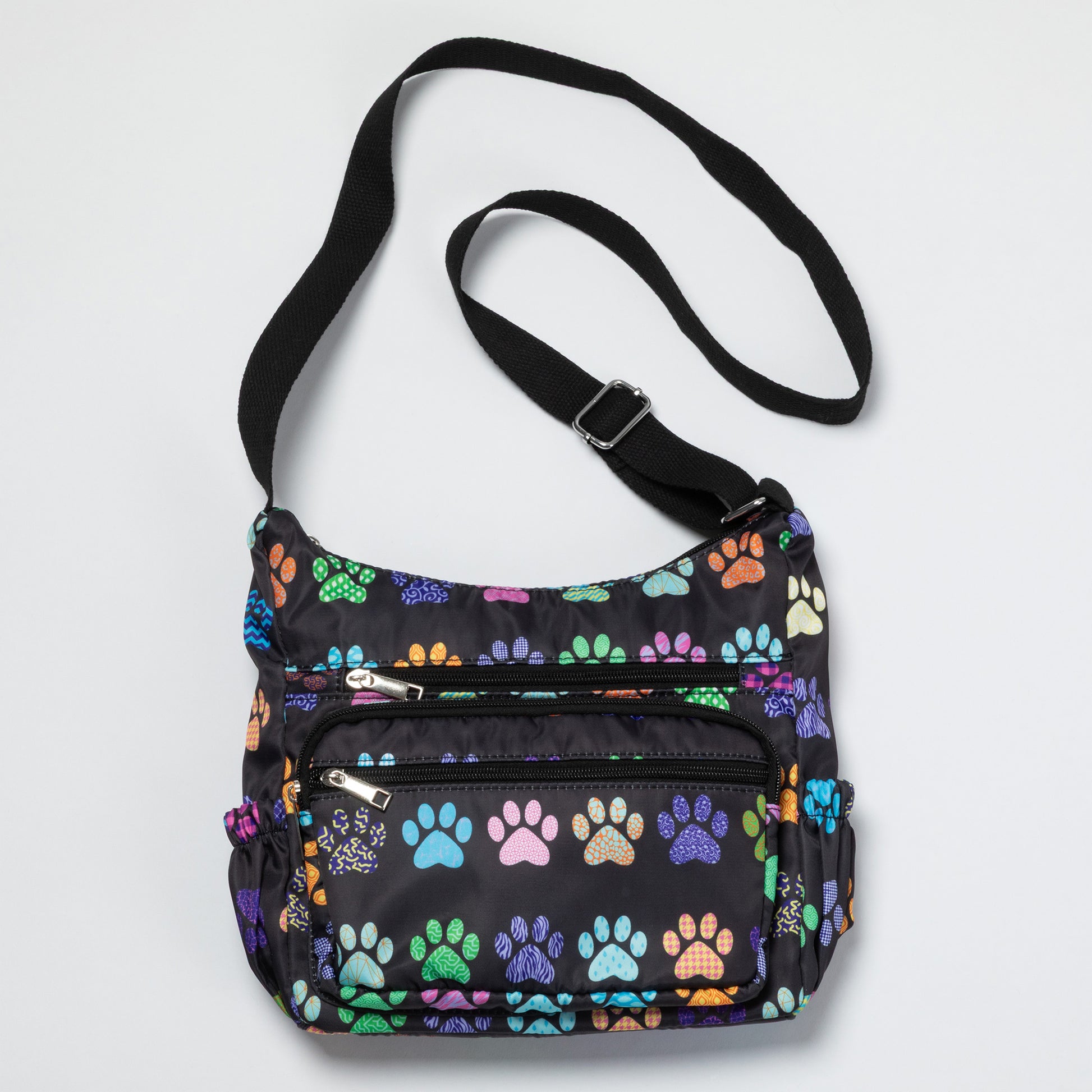 A black nylon crossbody bag featuring a colorful pattern of dog paw prints in various colors, with a black adjustable shoulder strap and silver zipper hardware.