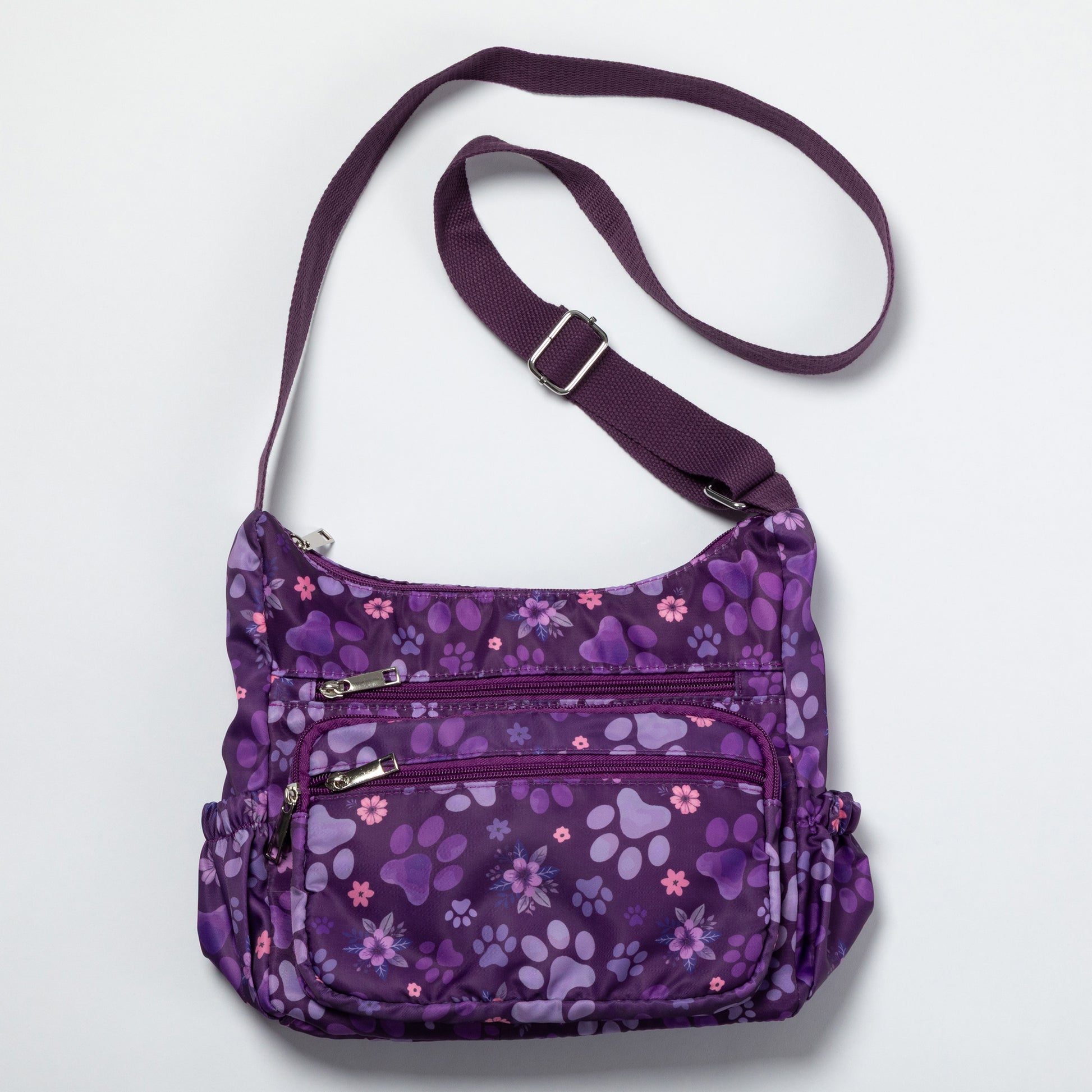 A purple crossbody bag with a floral and paw-print pattern, featuring multiple zippered pockets and a matching adjustable shoulder strap, shown on a light gray background.