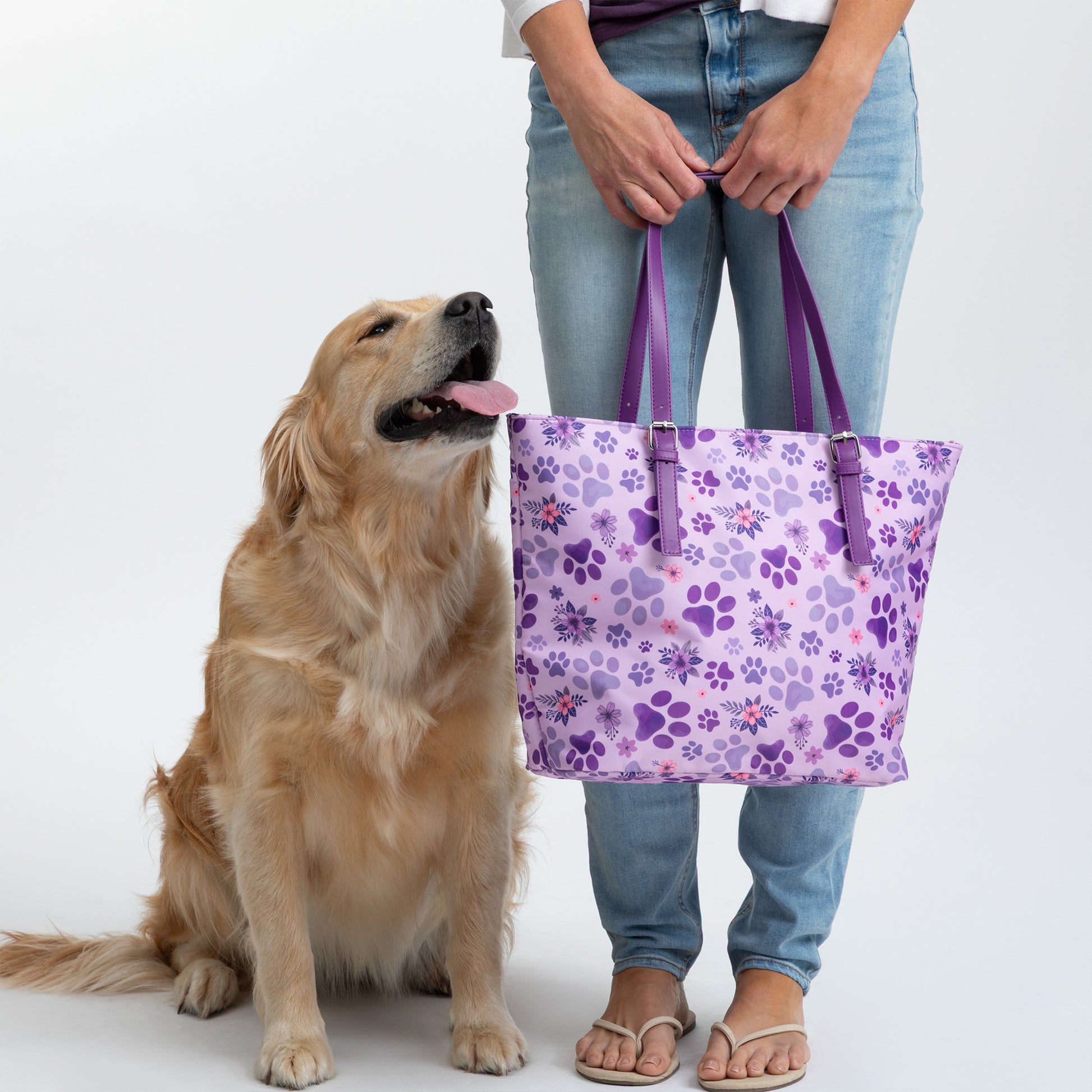 A person holds a purple floral tote bag beside a golden retriever.