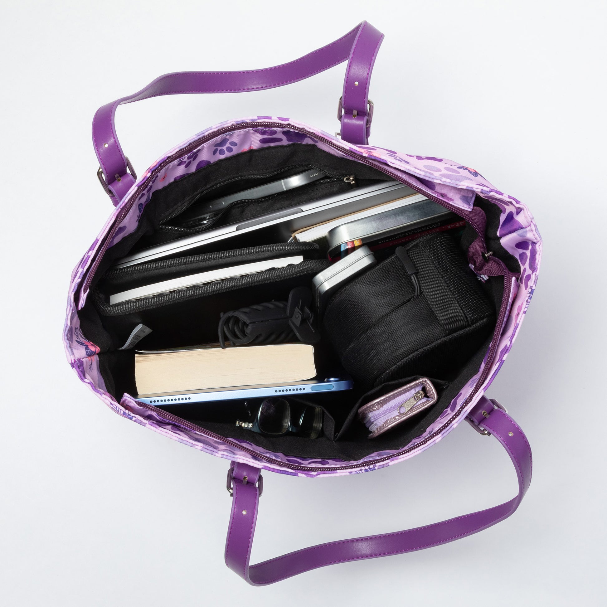A purple paw printed tote sitting open against a white background, showing the pockets inside and holding books, laptop, tablet, sunglasses etc.