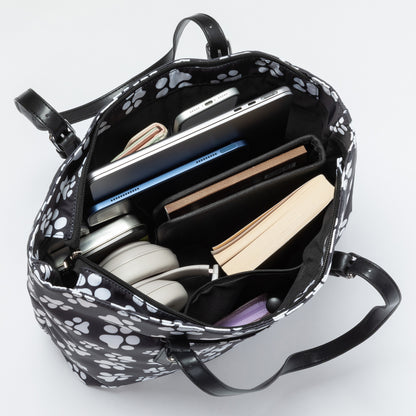 Black and white paw print tote bag shown open from above, filled with a laptop, notebooks, headphones, and other accessories, on a light gray background.