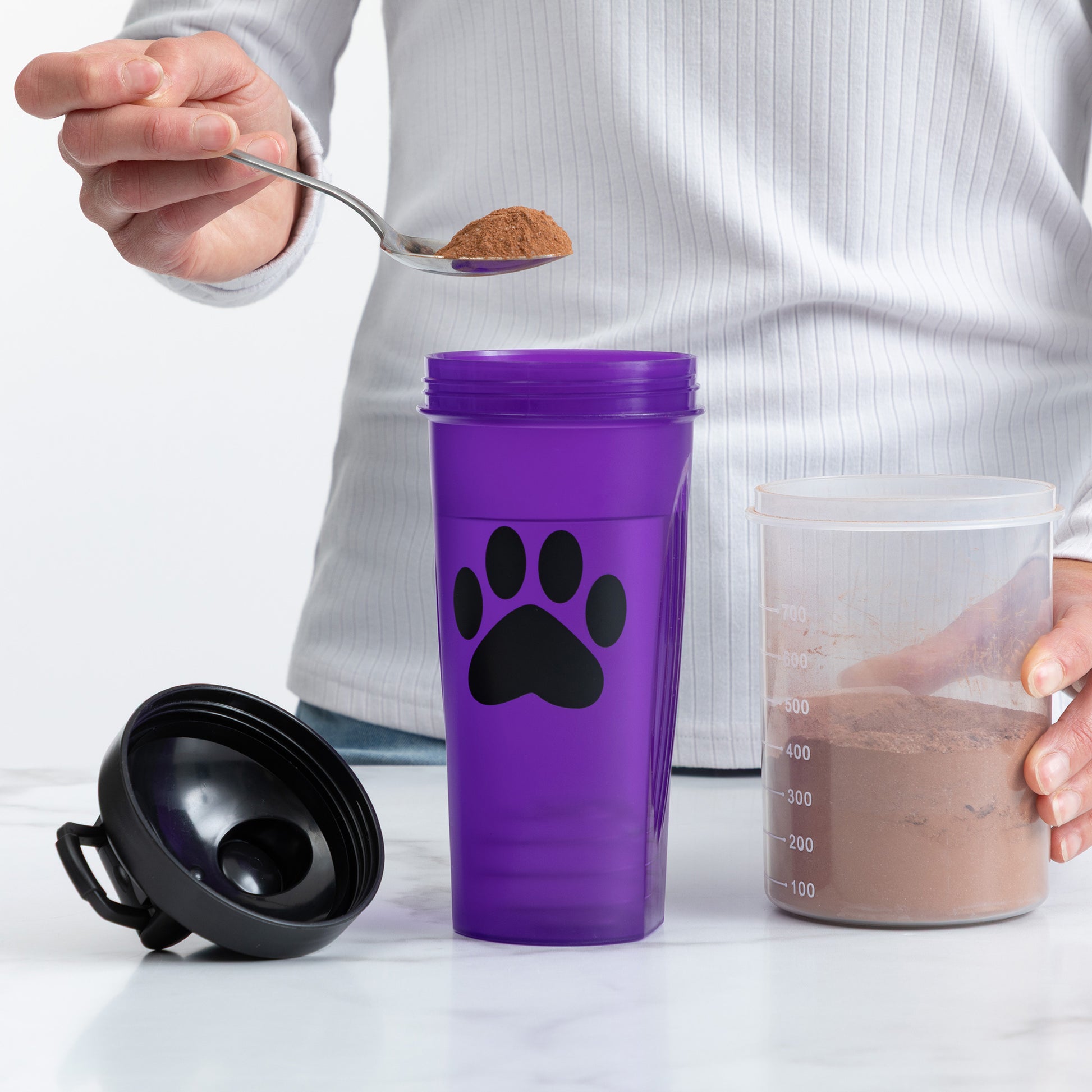 A person holds a spoon with brown powder above a purple shaker bottle with a black paw print, while holding a clear container filled with the mixed powder on a white marble surface.