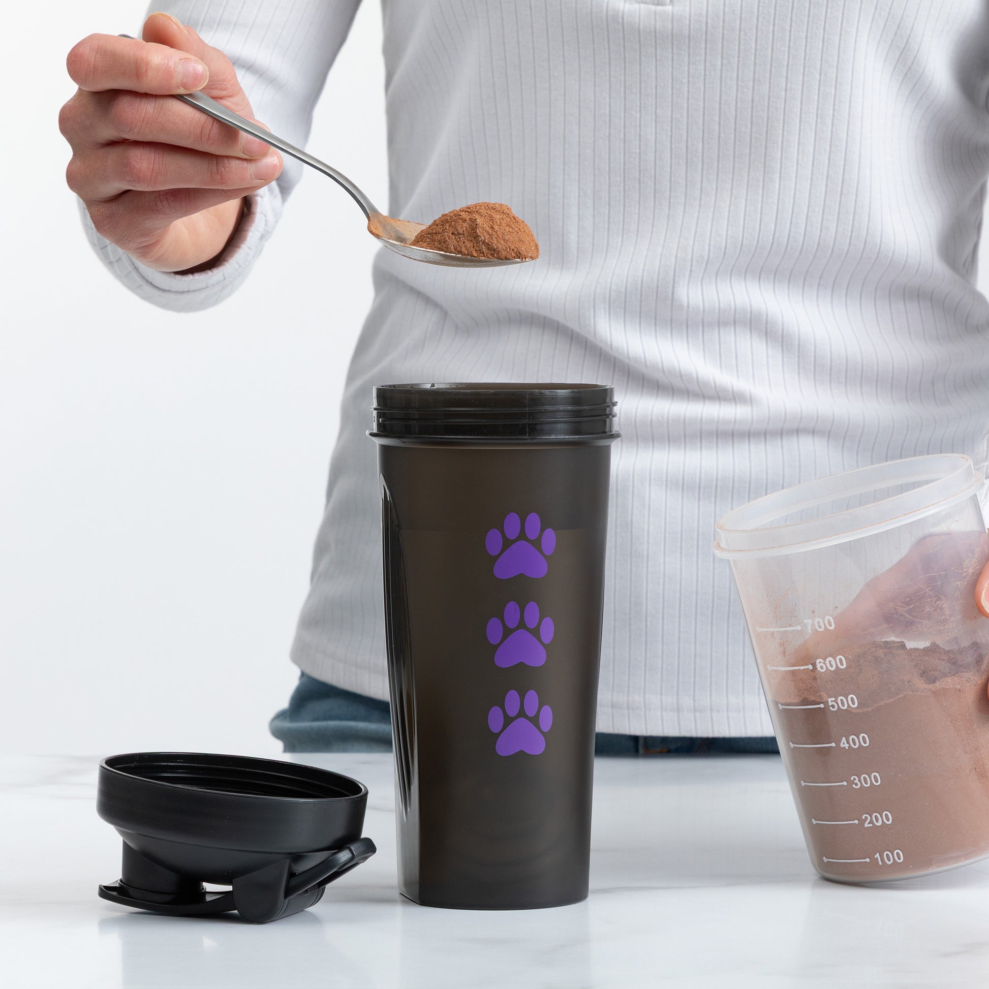 A person holds a spoon with brown powder above a dark gray travel tumbler with three purple paw prints. The tumbler lid is placed beside it, and a clear measuring cup containing the powder is visible on the right against a clean white background.