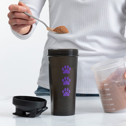 A person holds a spoon with brown powder above a dark gray travel tumbler with three purple paw prints. The tumbler lid is placed beside it, and a clear measuring cup containing the powder is visible on the right against a clean white background.