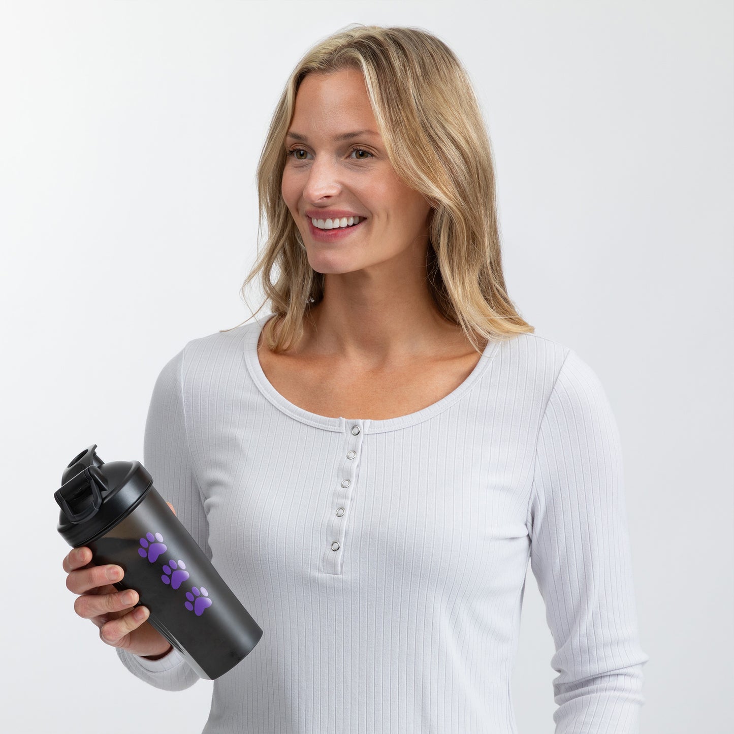 A smiling blonde woman wearing a white ribbed henley shirt holds a black shaker bottle with purple paw prints against a light gray studio background.