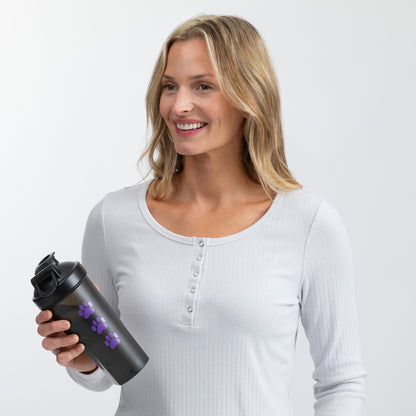 A smiling blonde woman wearing a white ribbed henley shirt holds a black shaker bottle with purple paw prints against a light gray studio background.