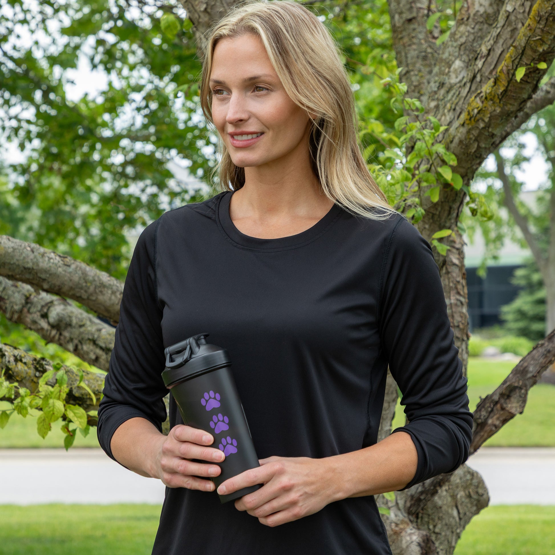 A woman standing outdoors in front of a tree, wearing a black long-sleeve athletic shirt and holding a black travel tumbler with purple paw print graphics.