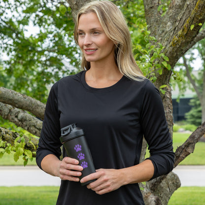 A woman standing outdoors in front of a tree, wearing a black long-sleeve athletic shirt and holding a black travel tumbler with purple paw print graphics.