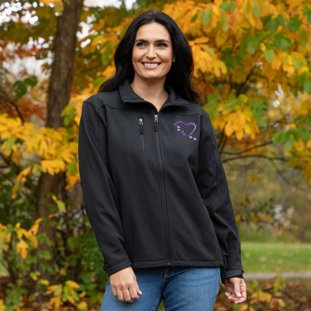 A woman wearing a black zip-up jacket with a purple heart graphic on the left chest, standing outdoors in front of autumn trees with yellow and green leaves.