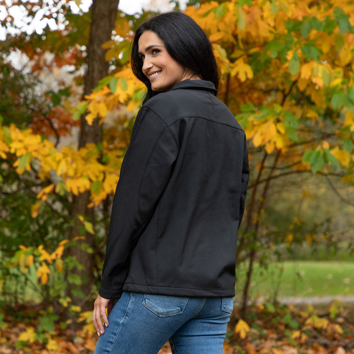 A woman wearing a black softshell jacket and blue jeans, photographed outdoors in a park with autumn trees in the background.