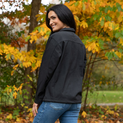 A woman wearing a black softshell jacket and blue jeans, photographed outdoors in a park with autumn trees in the background.