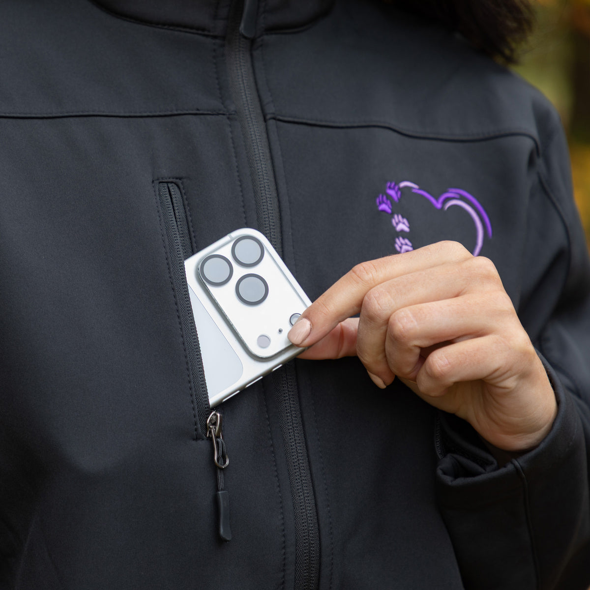 A close-up of a person wearing a black zip-up jacket with a purple heart-shaped paw print logo on the chest. The person is holding a silver smartphone in their hand, showing how the phone fits into a small zippered pocket on the jacket.