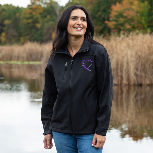A woman wearing a black zip-up jacket with a purple ribbon heart logo on the left chest, standing outdoors in front of a lake with reeds and trees in the background.