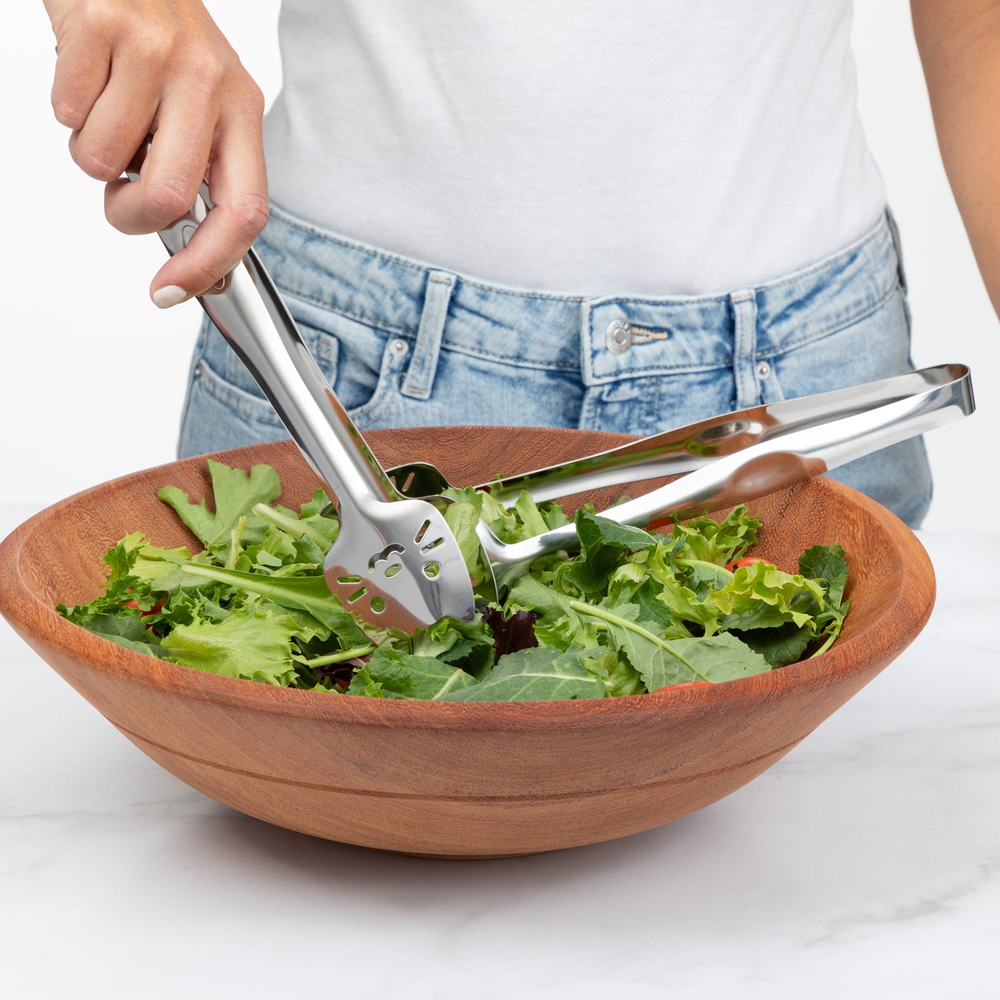 Person using stainless steel tongs to serve fresh salad from a wooden bowl.