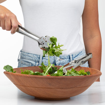 Person using stainless steel tongs to serve fresh salad from a wooden bowl.