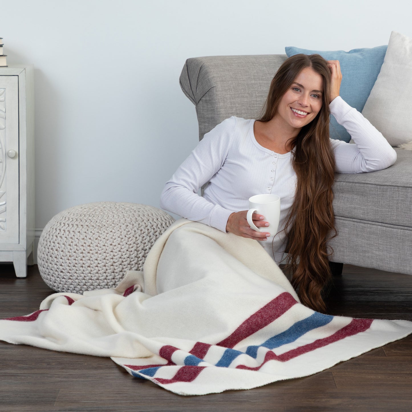 A woman sits on a gray armchair in a living room setting, wrapped in a cream-colored knit blanket with red and blue stripes. She is holding a white mug and wearing a white long-sleeve top.