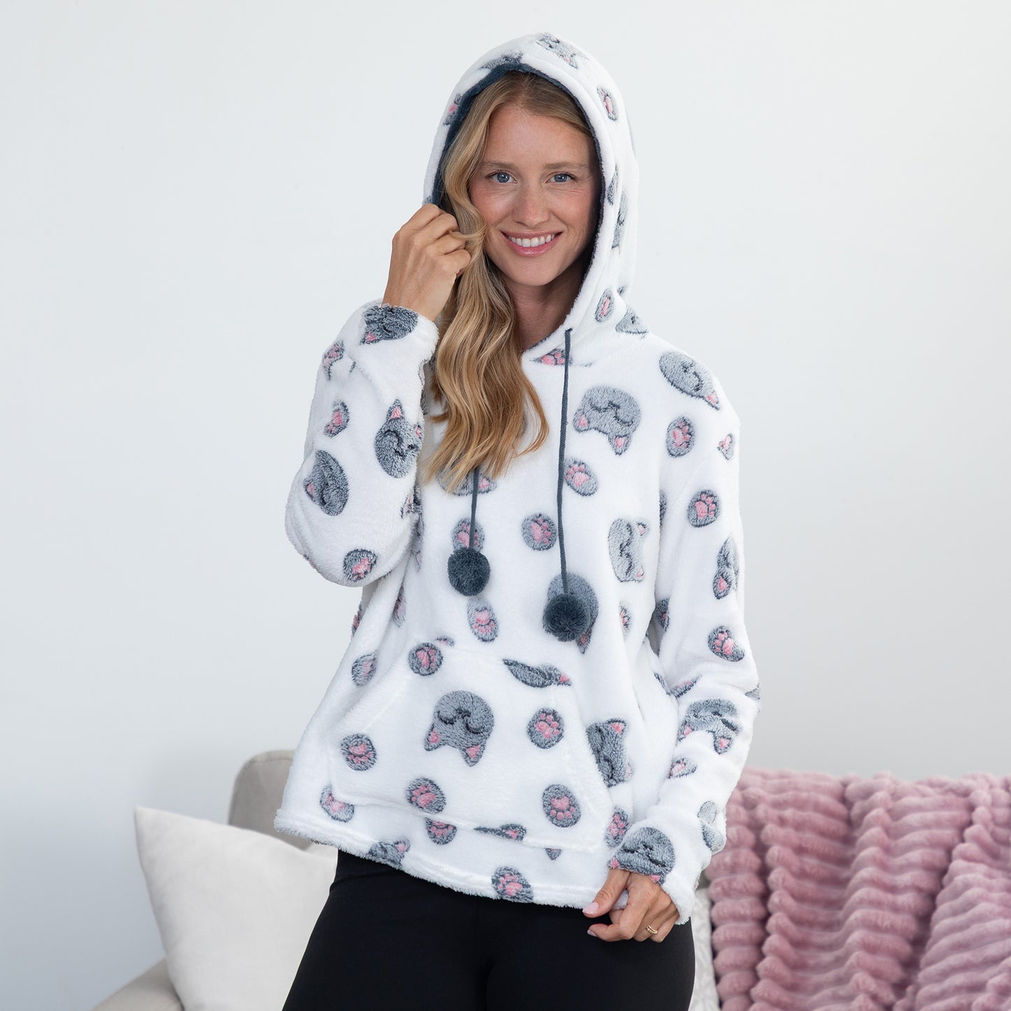 A woman wearing a white fleece hoodie with a gray cat print pattern, featuring black drawstrings with pom-poms, posing in a brightly lit room.
