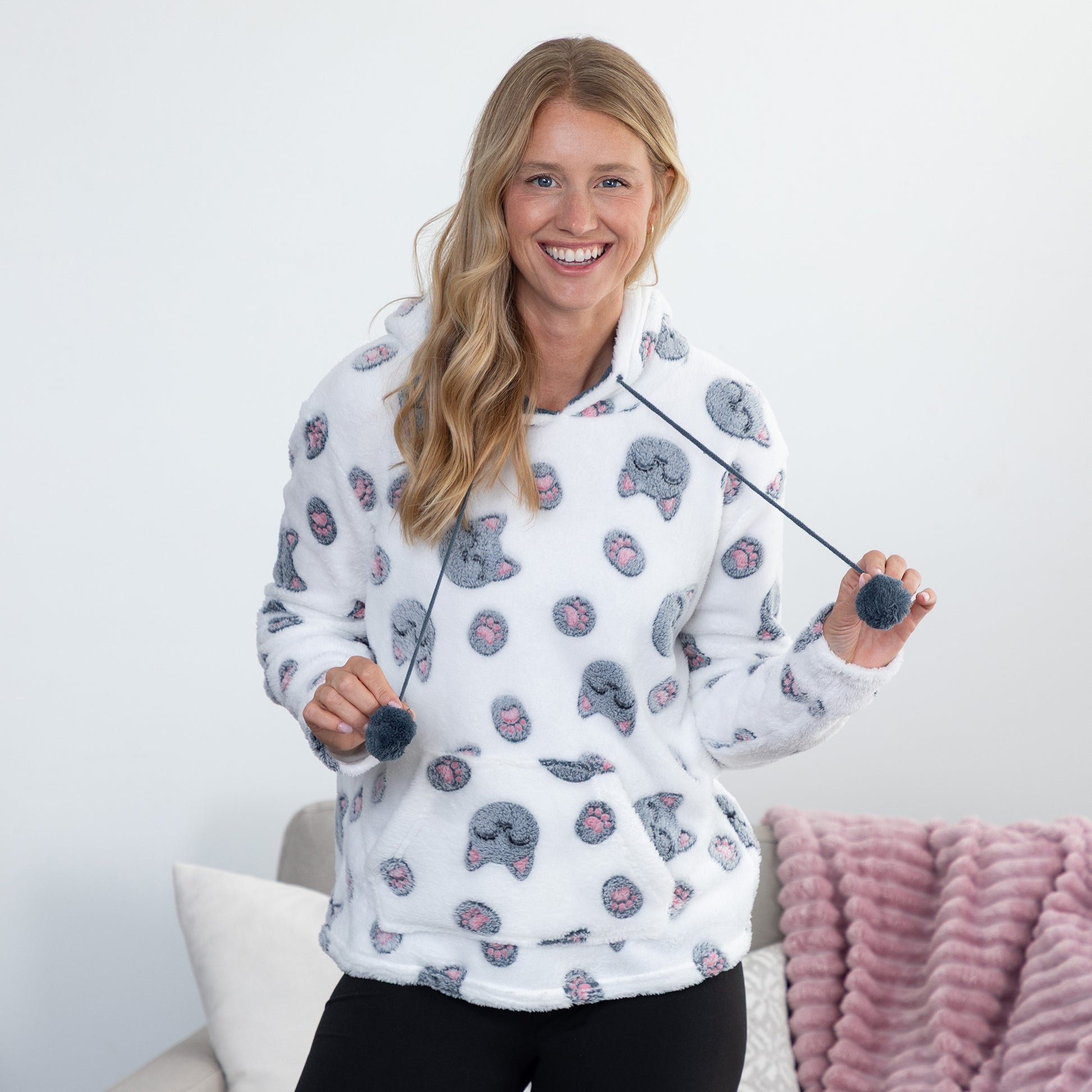 A smiling blonde woman wearing a white fleece hoodie with a gray cat print pattern, holding a toy with dark gray pom-poms in a brightly lit room.