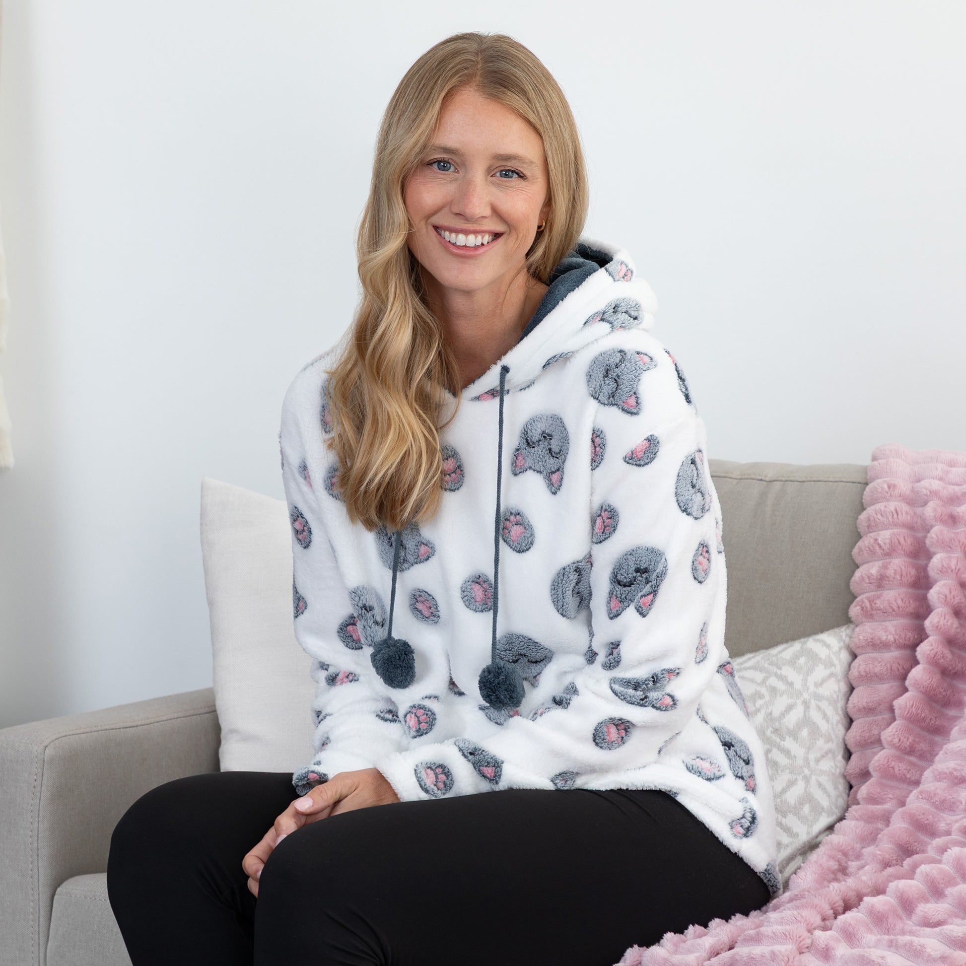 A smiling blonde woman sits on a light gray couch wearing a white hooded sweatshirt covered in a gray cat print pattern, paired with black pants. A pink textured throw blanket is draped over the couch beside her.