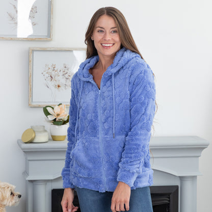 A smiling woman wearing a textured, plush light blue zip-up hoodie stands in front of a light gray fireplace mantel in a bright, modern room.