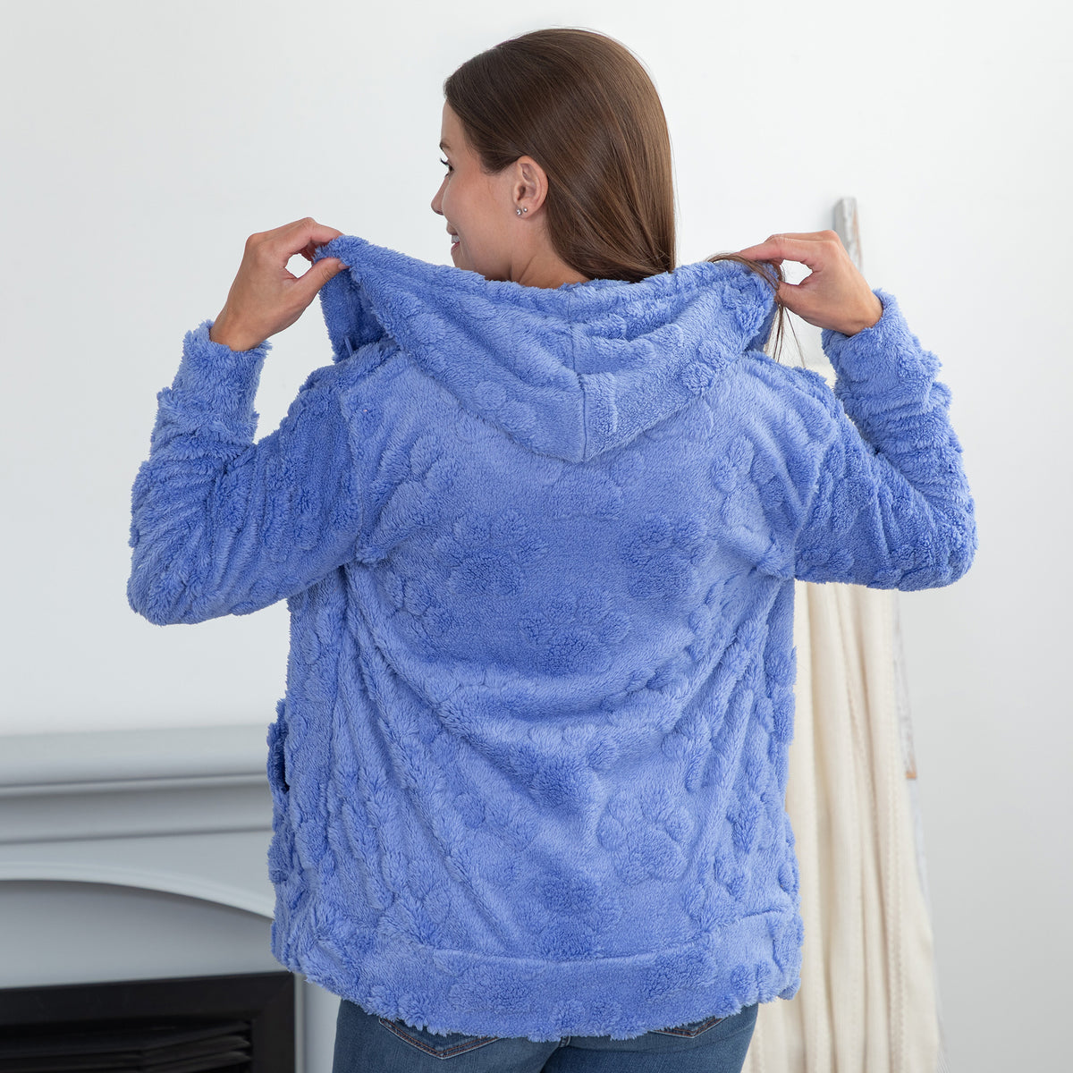 A woman viewed from behind wearing a textured, plush blue hooded sweatshirt, holding up the hood with both hands. 