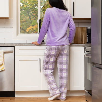 A woman stands with her back to the camera in a bright kitchen, wearing a lavender long-sleeve fleece top and matching plaid pajama pants with a small floral pattern.