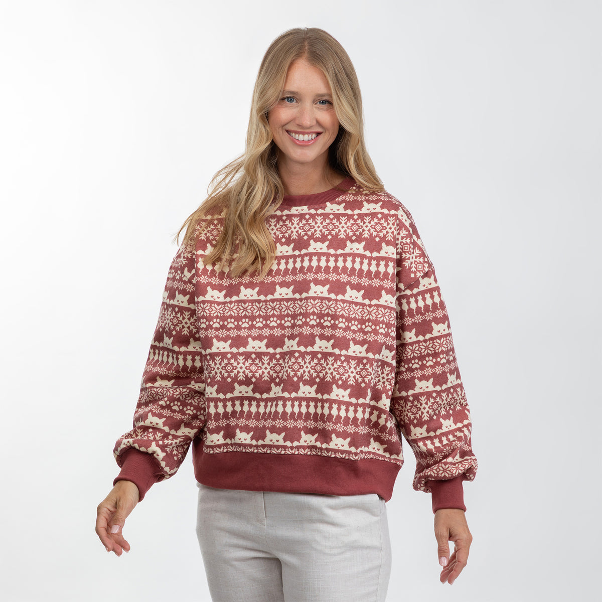 A woman modeling a long-sleeve crewneck sweatshirt with a festive fair isle pattern in rust red and cream, paired with light gray sweatpants against a plain white studio background.