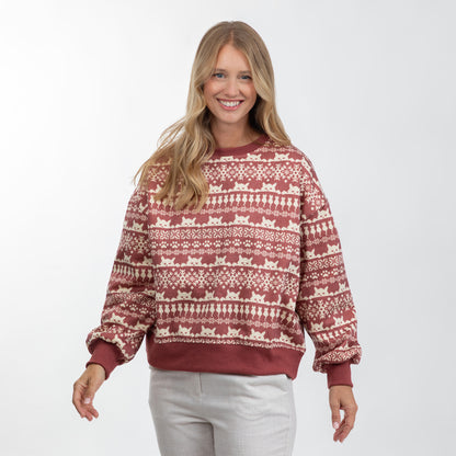 A woman modeling a long-sleeve crewneck sweatshirt with a festive fair isle pattern in rust red and cream, paired with light gray sweatpants against a plain white studio background.