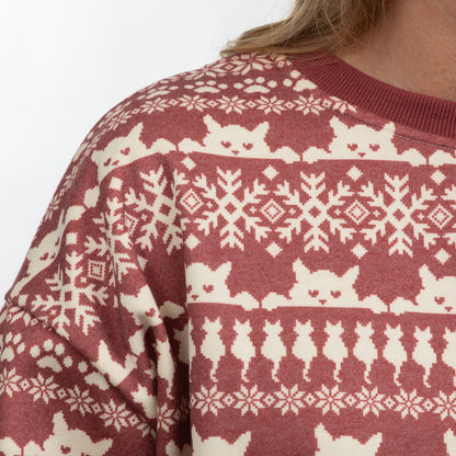 A close-up of a person wearing a festive crewneck sweater with a maroon and cream cat-themed Fair Isle pattern, including snowflakes, hearts, and cat silhouettes.
