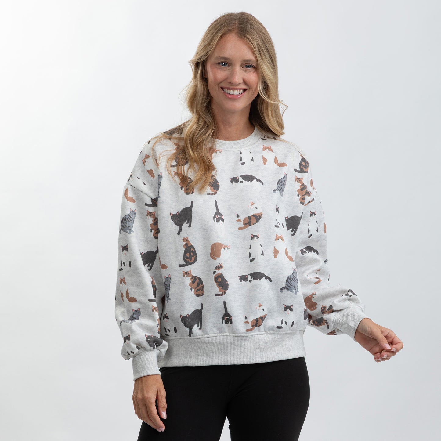 A woman modeling a light gray crewneck sweatshirt with an all-over print of various cat illustrations in black, brown, and white, paired with black pants against a light gray studio background.