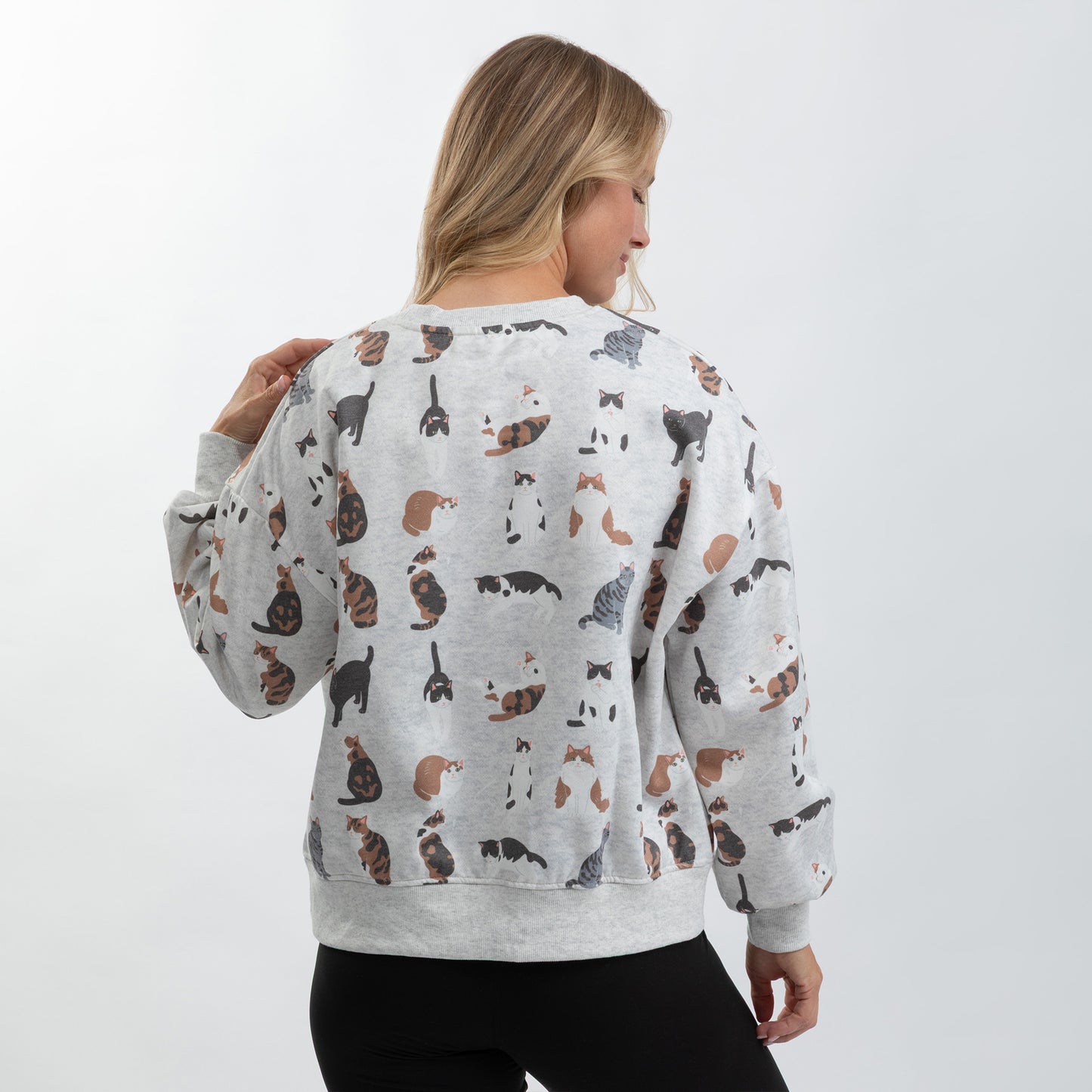 A woman viewed from the back wearing a light gray crewneck sweatshirt covered in a repeating print of various cat illustrations in black, brown, and white, paired with black pants against a plain white studio background.