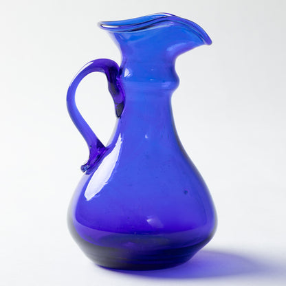A cobalt blue glass pitcher with a curved handle and a flared spout, shown on a clean white background.