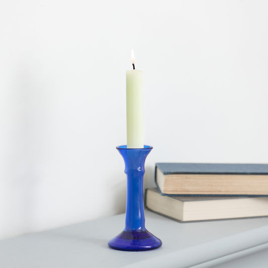 A lit cream-colored pillar candle standing in a cobalt blue glass candlestick holder, placed on a light gray surface with stacked books in the background against a white wall.