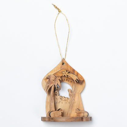 A handcrafted olive wood nativity ornament featuring a carved stable with a palm tree, star, baby Jesus, and animals, suspended by a natural jute string on a white background.