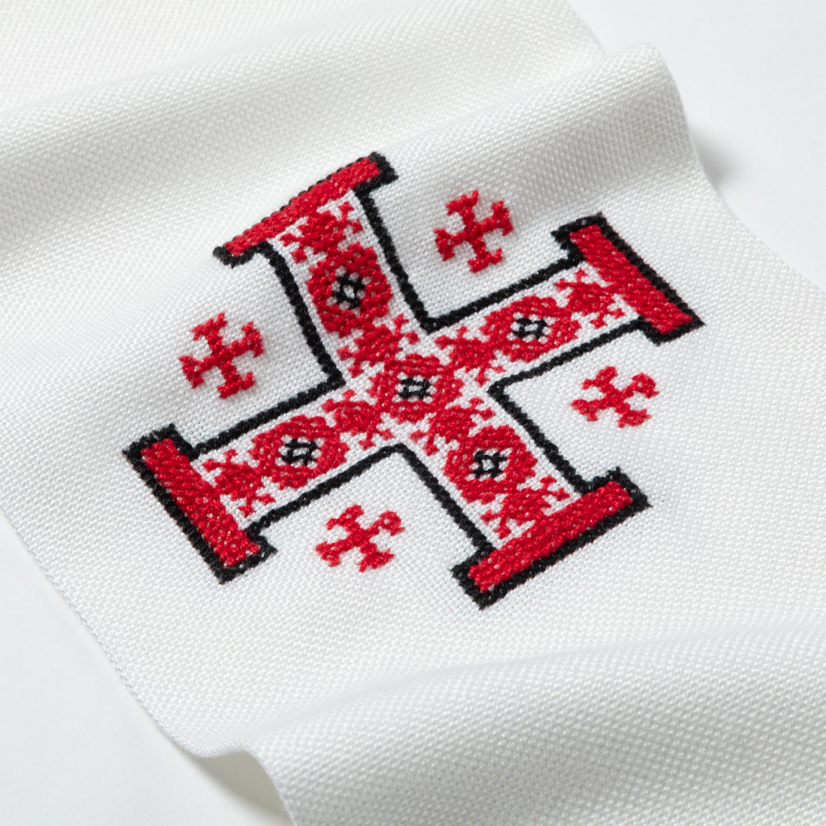 A close-up of a white knit fabric, likely a sock or scarf, featuring a red and black embroidered traditional cross design with geometric patterns.