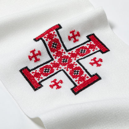 A close-up of a white knit fabric, likely a sock or scarf, featuring a red and black embroidered traditional cross design with geometric patterns.