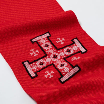A close-up of a red knit fabric scarf, featuring a large embroidered cross design in white and black.
