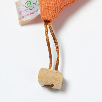 Close-up of an orange ribbed fabric doll with a wooden pom-pom on the tip of the hat, a wooden pom-pom on the bottom corner, and a small white tag attached