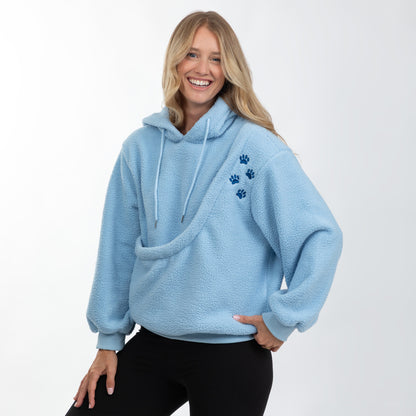 A smiling blonde woman wearing a light blue sherpa fleece hoodie with three dark blue paw prints on the left chest, paired with black pants, against a plain white studio background.