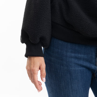 Close-up of a person wearing a black fleece pullover sweater with ribbed cuffs and hem, paired with blue jeans, against a plain white background.