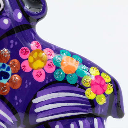 Close-up of a hand-painted ceramic dog magnet decorated with colorful floral patterns and glitter accents on a white background.