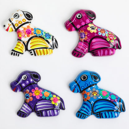 Four hand-painted ceramic dog figurines in vibrant colors, arranged in a 2x2 grid on a plain white background. Each dog features traditional Mexican folk art patterns, including floral motifs, stripes, and geometric shapes.