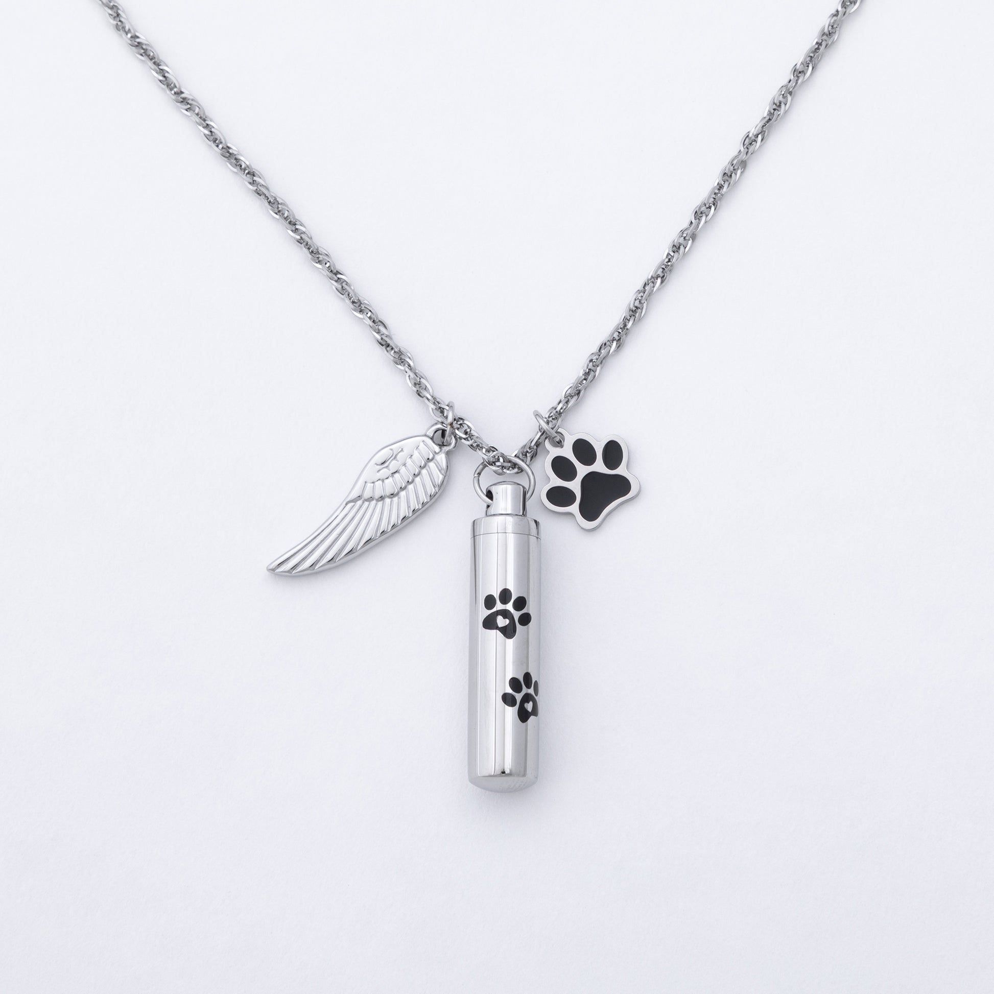 A silver-tone cremation urn necklace featuring a cylindrical vial pendant with black paw print engravings, an angel wing charm, and a black paw-shaped tag on a white background.