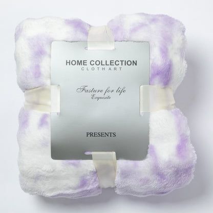 A folded plush throw blanket in white with soft lavender-purple tie-dye or watercolor-like patches, wrapped with cream ribbons and holding a silver hangtag that reads Home Collection Cloth Art.