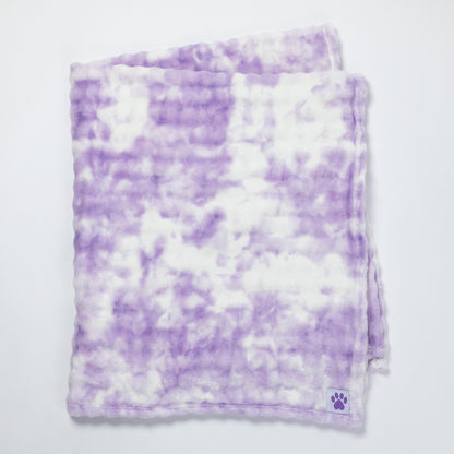 A folded plush blanket with a purple and white tie-dye pattern, featuring a small purple paw print logo in the bottom corner, set against a plain white background.